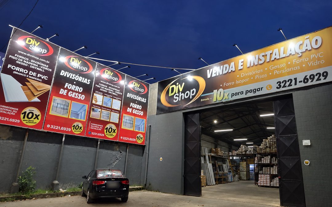 Fachada da loja da DivShop à noite, com as placas de forros e divisórias e o galpão aberto com materiais nas prateleiras